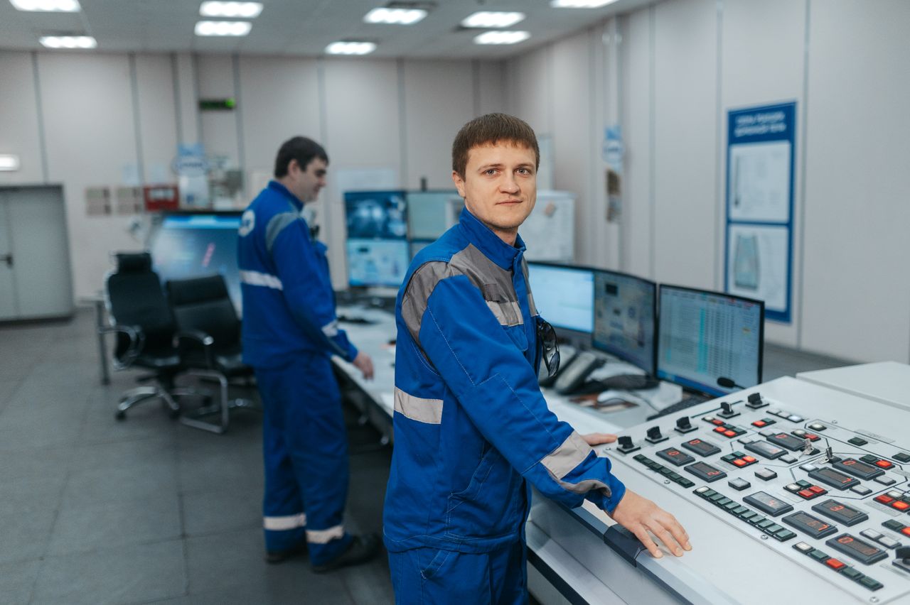 Home Two technicians operating machinery in a modern control room. Industrial setting with advanced equipment.